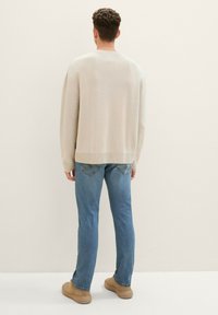 Suéter de punto beige con un corte relajado y cuello redondo, combinado con jeans de mezclilla azul claro y zapatos marrones. El fondo es de un tono neutro.