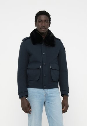 Marineblauwe jack met een nepbontkraag, voorknopsluiting en twee grote klepzakken. Draagt met lichtblauwe jeans met een gestructureerd ontwerp.