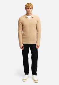 Beige strikket genser med polo-krage, lange ermer og et teksturert mønster, kombinert med svarte bukser og hvite sneakers.