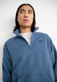 Pullover in pile blu chiaro con zip a metà collo, caratterizzato da un logo ricamato discreto e una finitura testurizzata. Maniche lunghe e tasche frontali.