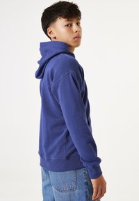 Sudadera con capucha azul de corte relajado, con puños y dobladillo acanalados, fabricada en un tejido suave y texturizado, y con un bolsillo delantero, combinada con jeans de mezclilla claros.