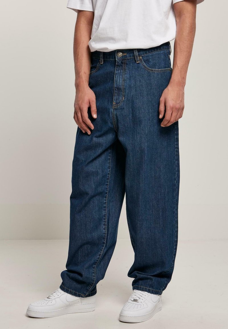 Jean en denim de couleur bleu foncé, coupe ample, avec braguette à boutons, poches avant, et baskets blanches. Texture lisse avec des coutures minimales.