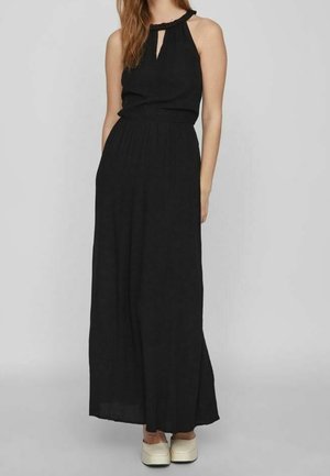 Kobieta ubraną w czarną, bezrękawnikową suknię maxi z dekoltem halter oraz białe buty na platformie, stojąca na tle prostego, jasnoszarego tła.