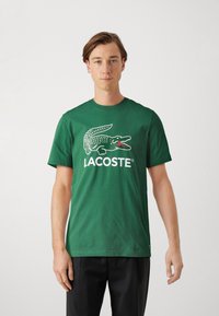 Žalias medvilninis marškinėliai su balta krokodilo logotipu ir raudonomis akcentais, o žemiau logotipo - ryškiai raudonas tekstas "LACOSTE". Trumpomis rankovėmis, apvaliu kaklu.