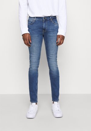 Mand iført tætsiddende blå jeans, hvid langærmet skjorte og hvide sneakers, stående mod en ensfarvet lys baggrund.