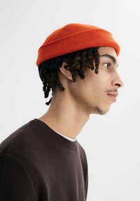 Oranje gebreide beanie met een blauwe elastische band, scheef gedragen. De achtergrond is effen wit, wat de kleurcontrasten en texturen van de muts benadrukt.