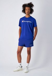 Mėlynas "Champion" marškinėlių su logotipu komplektas su mėlynais šortais, baltais sportbačiais ir raštuotomis kojinėmis, stovintis ant neutralaus fono.