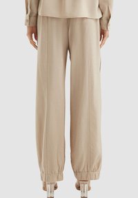 Beige wijde broek met een gladde textuur, elastische boorden en zijzakken, gedragen met een losse bijpassende top, gezien vanuit de achterkant.
