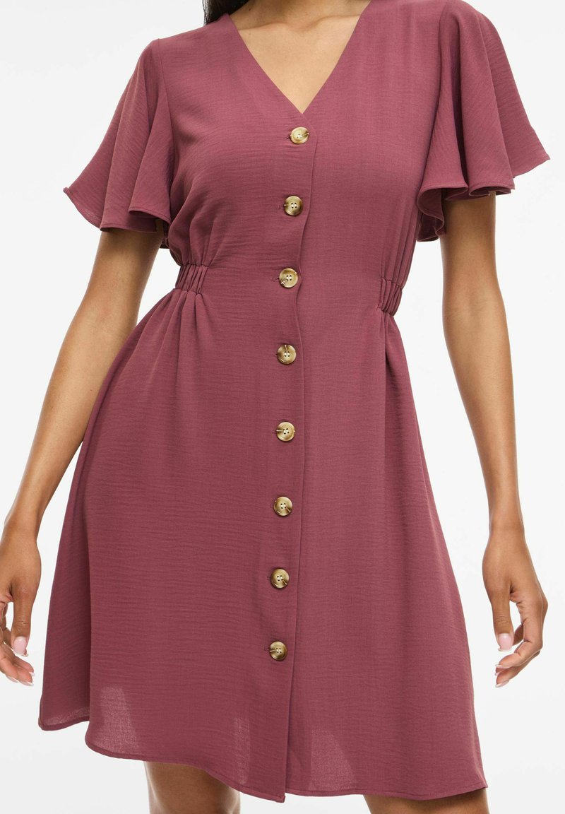 Femme portant une robe couleur vieux rose mi-longue, manches courtes, col en V et rangée de boutons marron sur le devant, taille cintrée, et manches flottantes.