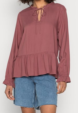 Femme portant un chemisier à manches longues couleur vieux rose avec un ourlet et des poignets volantés, associé à un short en jean bleu clair à hauteur des genoux.