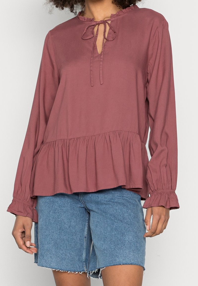 Femme portant un chemisier à manches longues couleur vieux rose avec un ourlet et des poignets volantés, associé à un short en jean bleu clair à hauteur des genoux.