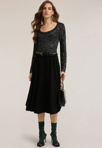 Czarna bluza z długim rękawem z błyszczącym wykończeniem, czarna spódnica typu A, opleciony czarny pasek, zielone teksturowane skarpetki i czarne sandały na szpilce z paskami.