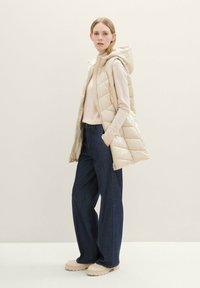 Beige Daunenweste mit Kapuze, gestepptes Muster, getragen über einem hellrosa Sweater und dunkelblausweiten Hosen, kombiniert mit beigen Plateauschuhen.
