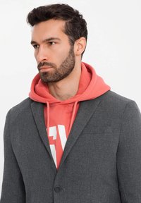 Grauer Blazer über einem korallenfarbenen Hoodie mit weißen Grafiken, ausgestattet mit einem Zwei-Knopf-Verschluss, Reverskragen und einem strukturierten Stoff.