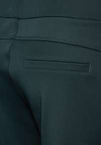 Mörkgröna byxor med en slank design, slät textur och en enda bakficka med räta sömmar.