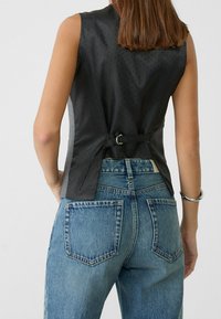 Czarny top bez rękawów z teksturowanym wzorem, z tylnym zapięciem na klamrę, w zestawieniu z jasnoniebieskimi, wysokim stanem dżinsami z kieszeniami.