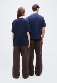 Δύο άτομα με την πλάτη στην κάμερα, φορούν μπλε ναυτικό t-shirt και καφέ παντελόνια με φαρδιά πόδια, σε απλό ανοιχτό φόντο.