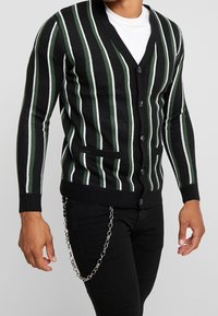 Cárdigan negro con rayas verticales en verde y blanco, diseño de cuello en V, siete botones y dos bolsillos frontales. Lleva puesto sobre una camisa blanca.