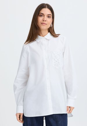 Woman with long brown hair wears a loose white button-up shirt with a cutout pattern pocket and dark blue jeans, standing against a plain background.