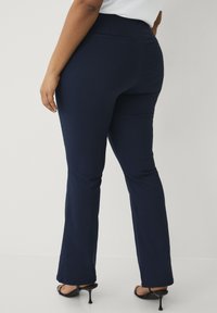 Navyblauwe skinny pants met een gladde textuur, uitlopende pijpen en een detail van een achterzak. Gemaakt met zwarte hoge hakken.