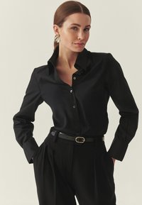 Fekete, gombos ing elegáns kialakít ással, szabott illeszkedéssel és hajtóka gallérral. Hosszú ujjakkal és fekete nadrággal, arany díszítésű övvel kiegészítve.