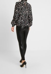 Blusa nera a fiori con maniche a sbuffo e dettaglio a occhiello sul retro, abbinata a leggings neri lucidi e scarpe nere con tacco alto.