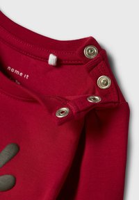 Rode katoenen blouse met een ronde halslijn, voorzien van zilveren drukknoopsluitingen op de schouder. Voorzien van een grijs gedrukt ontwerp.