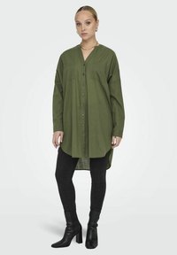 Chemise boutonnière vert kaki avec un col en V, deux poches poitrine et un ourlet arrière plus long, associée à un jean slim noir et des bottines pointues.