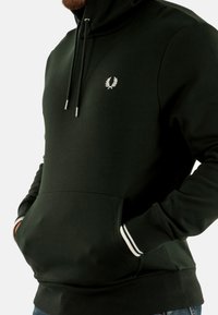 Sudadera con capucha de color verde oscuro, con cordones ajustables, bolsillo frontal y acentos a rayas blancas en los puños. Logo en el pecho.