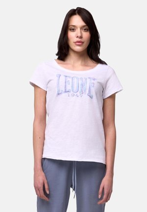 Femme aux cheveux foncés portant un t-shirt blanc « LEONE 1947 » et un pantalon bleu à cordon, debout devant un fond clair uni.