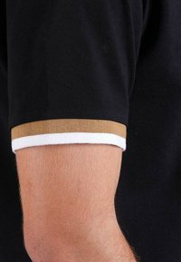 Polo noir en tissu texturé, avec un poignet rayé marron et blanc à la manche. Gros plan sur un détail de bras.