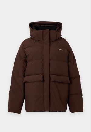 Femme portant une doudoune marron foncé avec une capuche, un pull à col roulé beige, un jean évasé bleu et des chaussures foncées, debout devant un fond uni.