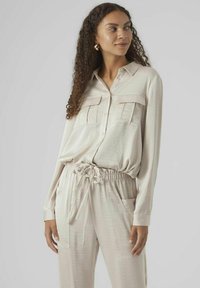 Chemise beige clair à manches longues avec deux poches poitrine, associée à un pantalon à cordon de serrage assorti. Texture lisse avec une coupe décontractée.