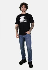 T-shirt in cotone nero con grande logo bianco "STARTER" e design a stella, abbinato a jeans blu e scarpe da ginnastica nere. Il modello sta in piedi rivolto in avanti.