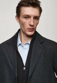 Grauer Wollmantel über einem schwarzen Reißverschluss-Pullover und einem hellblauen Hemd. Der Mantel verfügt über einen Reverskragen und einen Reißverschluss, der die übereinanderliegenden Texturen zur Geltung bringt.