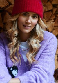 Levanduľový pletený sveter s čiernobielym motívom mačky, červená rebrovaná čiapka a farebný náhrdelník z korálikov. Mäkká textúra, voľný strih.