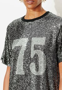 T-shirt noir à sequins avec un grand imprimé argenté "75", manches courtes à franges et un col rond noir côtelé.