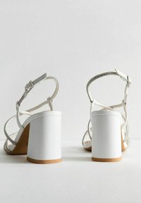 Sandali bianchi con tacco a blocco, dotati di fasce metalliche e una cinghia alla caviglia textured, che mettono in mostra un design minimalista.