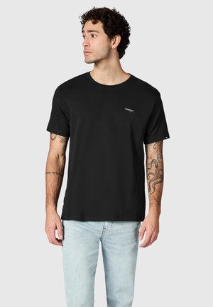 Junger Mann mit lockigem Haar, trägt ein einfaches schwarzes T-Shirt und hellblaue Jeans, schaut nach links und zeigt Tätowierungen auf beiden Unterarmen.