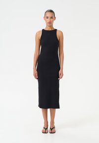 Abito midi nero a coste con design senza maniche, scollatura tonda e spacchi laterali per facilità di movimento. Indossato con sandali neri con suola piatta. Tessuto liscio.