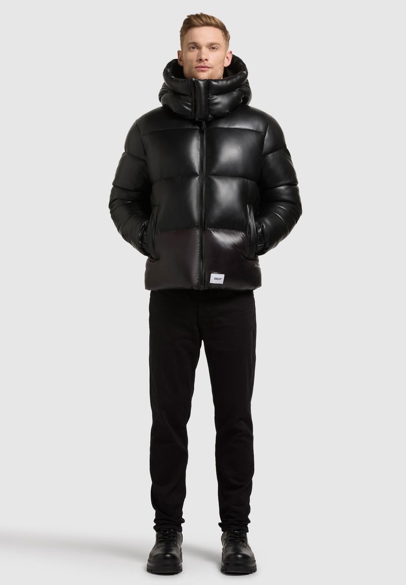 Black puffer jacket featuring a high collar, zip closure, and large quilted sections. Paired with black pants and sturdy black boots.