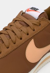 Smeđi tenisice s teksturiranim kožnim i platnenim gornjim dijelom, sa svijetlosmeđim Nike značkom, smešim vezicama i bijelim oznakom na jeziku.
