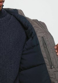 Kurtka puchowa o szarym zewnętrznym materiale i ciemnoniebieskiej podszewce, z wysuwaną kieszenią i prążkowanym swetrem w kolorze granatowym pod spodem. Gładka faktura.