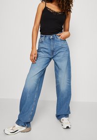 Szerokie niebieskie dżinsy z wysokim stanem, wykonane z denimu. Noszone z czarnym topem na ramiączkach z koronkową lamówką oraz białymi trampkami z detalami.