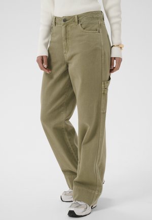 Pantalon ample vert olive avec poche utilitaire sur le côté, porté avec des baskets blanches et un haut crème à manches longues côtelé, modèle montré de la taille aux pieds.