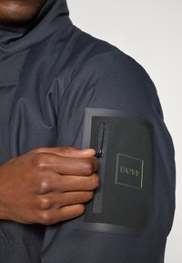 Jacket preta texturizada com um design elegante, apresentando um bolso com fecho de correr no braço. O bolso tem um destaque com um logótipo verde e hardware moderno.