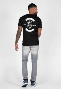Sort bomulds t-shirt med en hvid grafisk design på ryggen, som viser en karakter og brandtekst; parret med lysegrå jeans og sneakers.