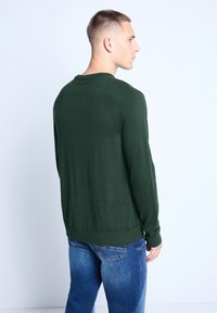Pull vert foncé en maille légère, à col rond et avec des poignets côtelés. Porté avec un jean en denim bleu.