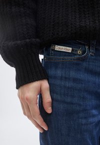 Suéter negro de punto con puños acanalados, combinado con jeans de mezclilla azul oscuro que tienen una etiqueta blanca y roja de "Calvin Klein" en el bolsillo.