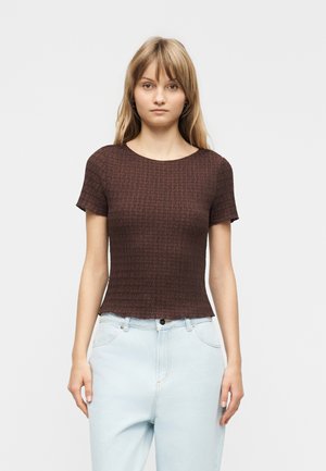 Junge Frau mit glatten blonden Haaren, die ein strukturiertes braunes Kurzarmoberteil und hellblaue hoch taillierte Jeans trägt, vor weißem Hintergrund stehend.